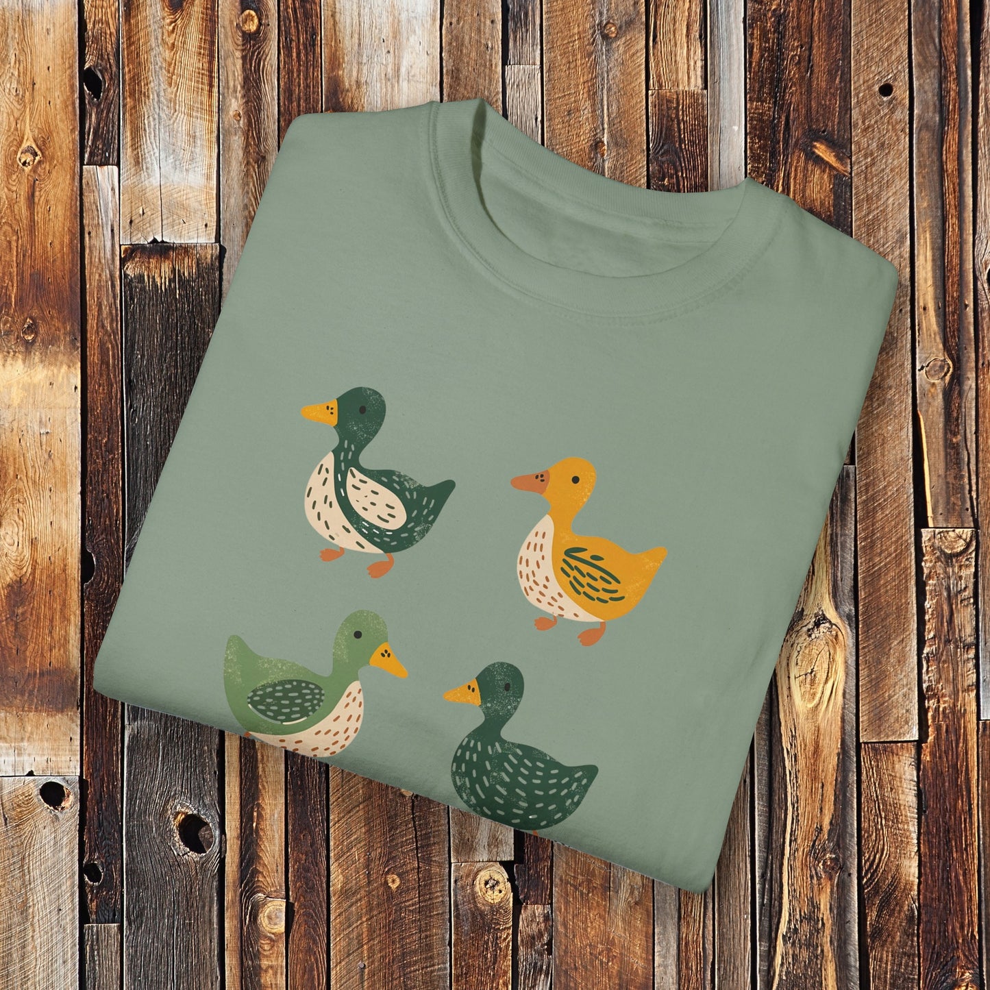 Light green t-shirt featuring a cute illustration of four cartoon ducks in earthy tones of green, yellow, and beige. Each duck has a unique color and pattern, arranged in a simple, playful style, adding a charming touch to the design.