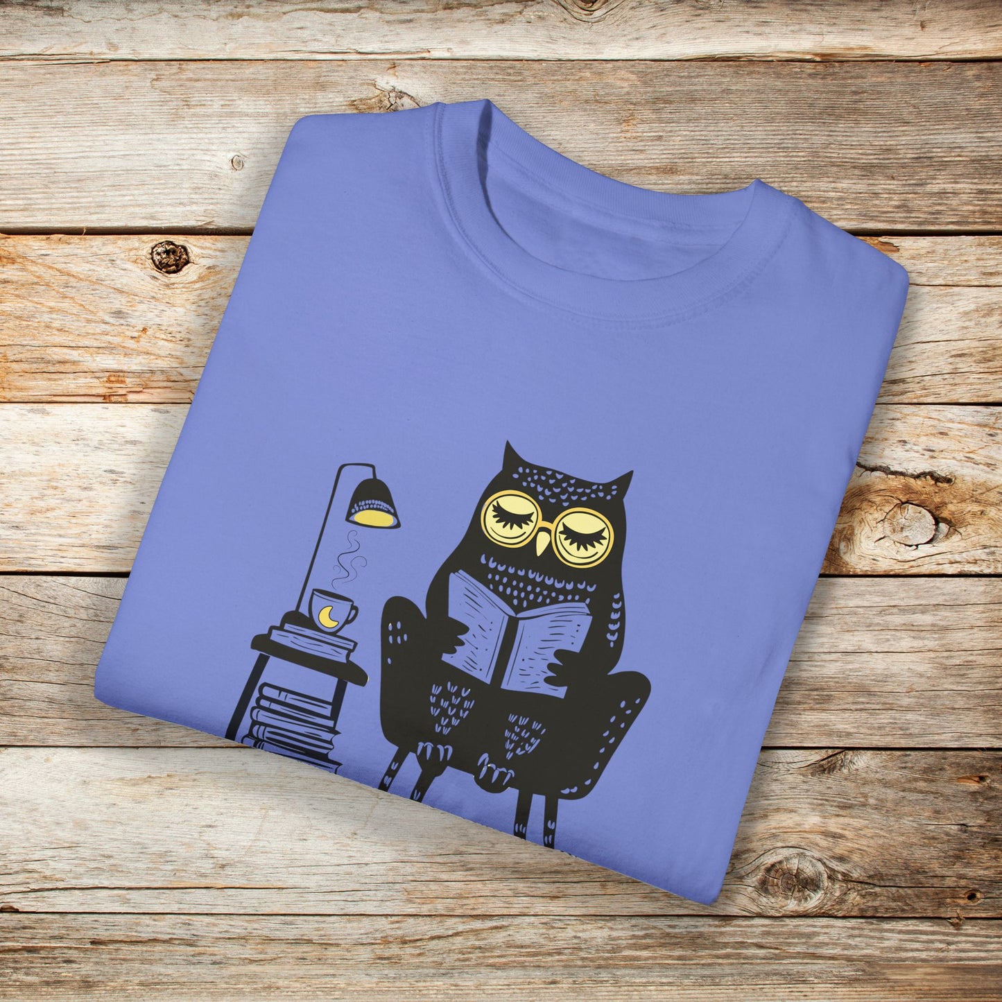 Blue t-shirt featuring a line art drawing of an owl with large yellow eyes, sitting on a chair, reading a book. There is a stack of books beside her, and below, the text 'Night Owl' is written in bold letters, adding a playful touch to the design.