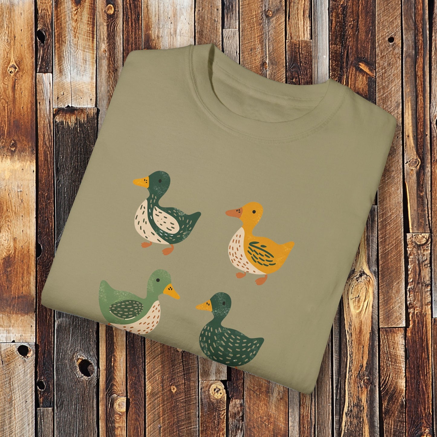 Khaki colored t-shirt featuring a cute illustration of four cartoon ducks in earthy tones of green, yellow, and beige. Each duck has a unique color and pattern, arranged in a simple, playful style, adding a charming touch to the design.