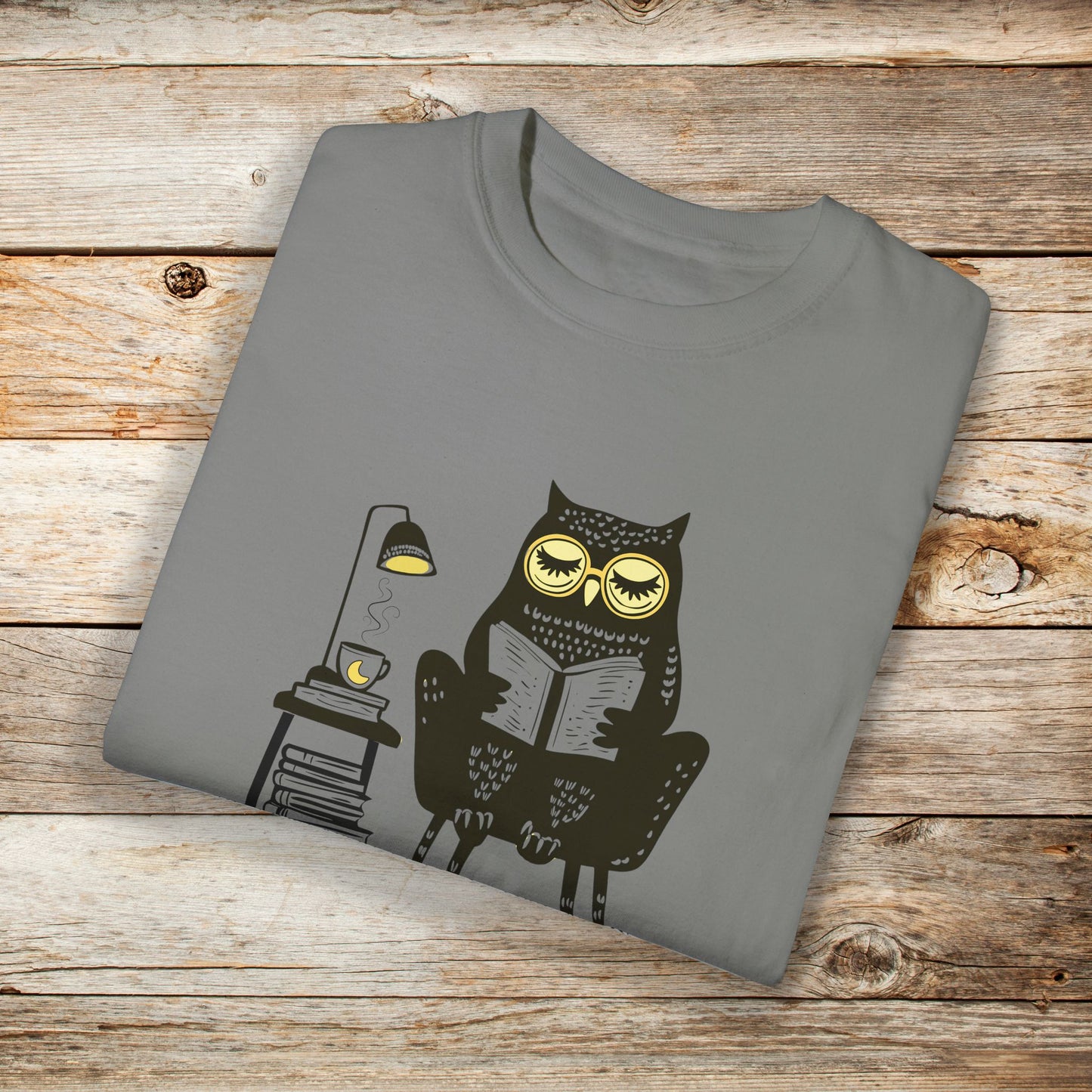 Light gray t-shirt featuring a line art drawing of an owl with large yellow eyes, sitting on a chair, reading a book. There is a stack of books beside her, and below, the text Night Owl is written in bold letters, adding a playful touch to the design.