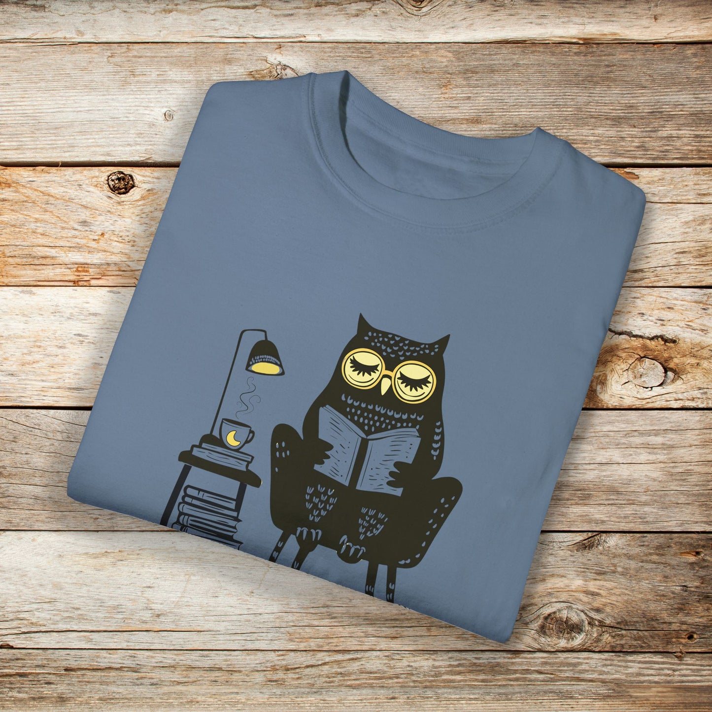Blue t-shirt featuring a line art drawing of an owl with large yellow eyes, sitting on a chair, reading a book. There is a stack of books beside her, and below, the text Night Owl is written in bold letters, adding a playful touch to the design.