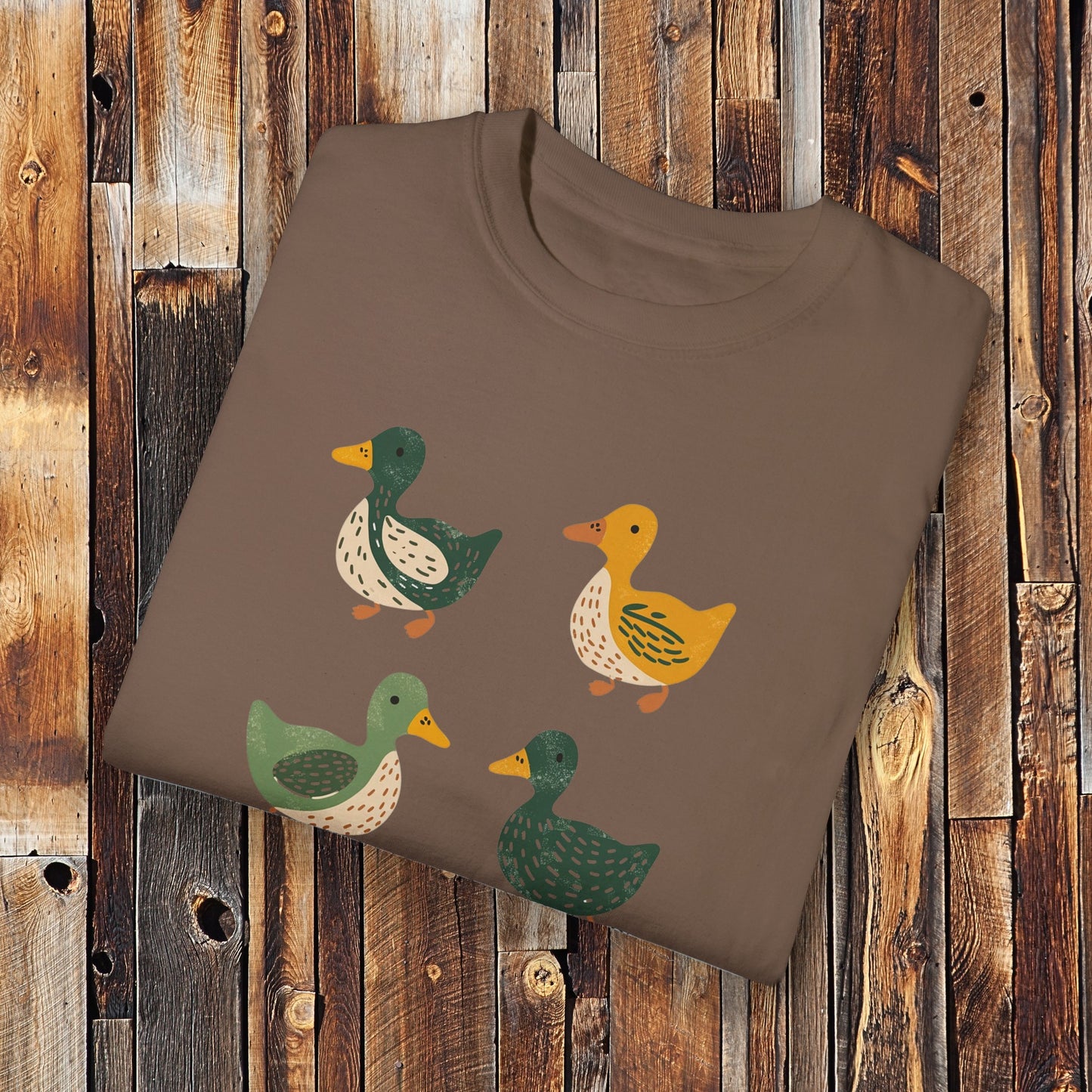 Espresso colored t-shirt featuring a cute illustration of four cartoon ducks in earthy tones of green, yellow, and beige. Each duck has a unique color and pattern, arranged in a simple, playful style, adding a charming touch to the design.