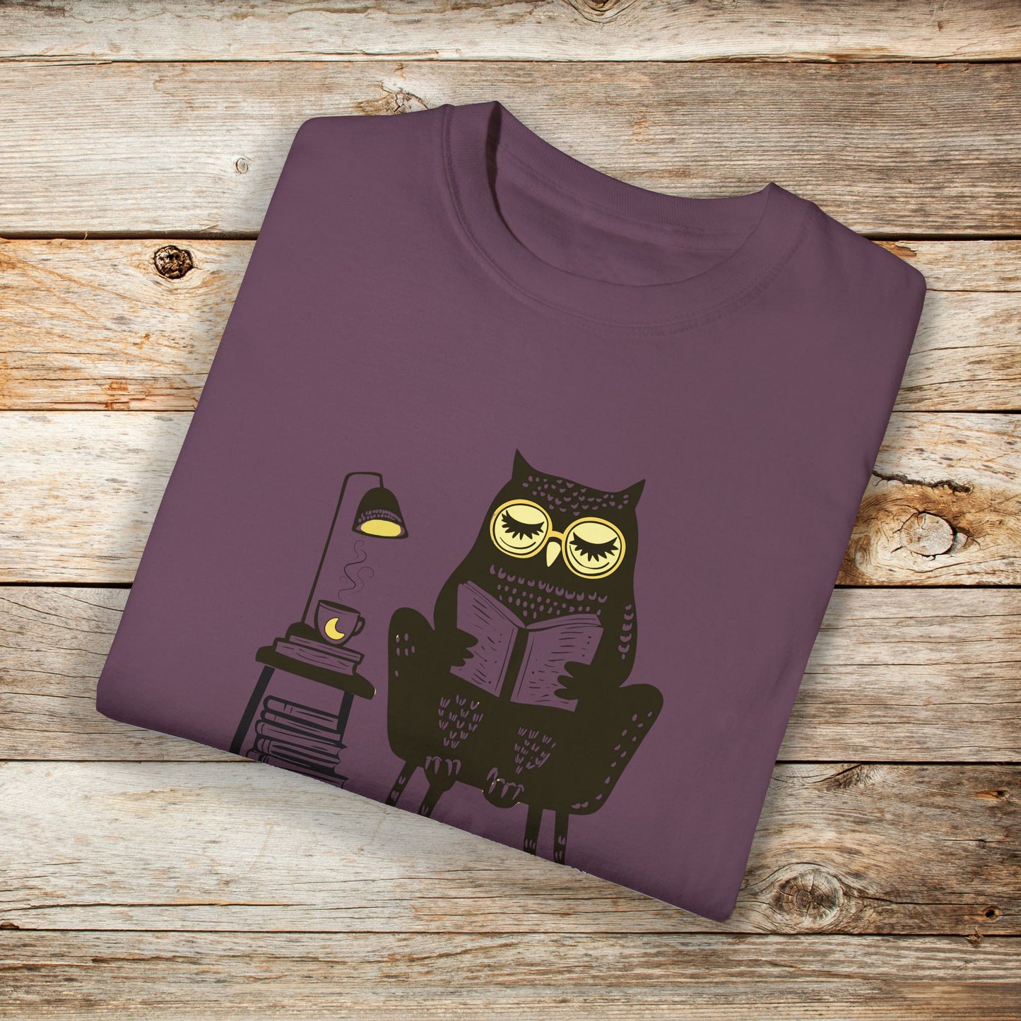 Purple t-shirt featuring a line art drawing of an owl with large yellow eyes, sitting on a chair, reading a book. There is a stack of books beside her, and below, the text Night Owl is written in bold letters, adding a playful touch to the design.