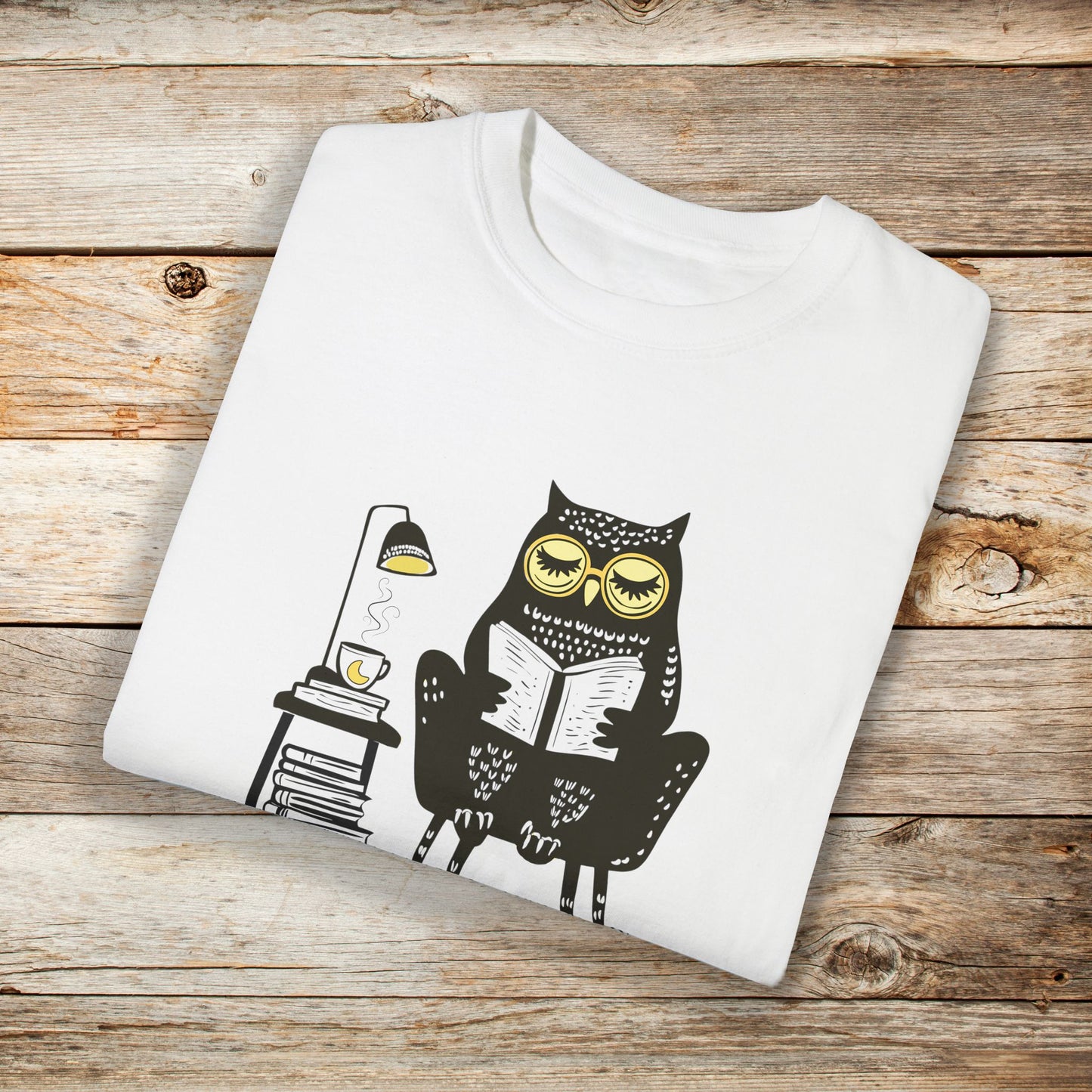 White t-shirt featuring a line art drawing of an owl with large yellow eyes, sitting on a chair, reading a book. There is a stack of books beside her, and below, the text 'Night Owl' is written in bold letters, adding a playful touch to the design.