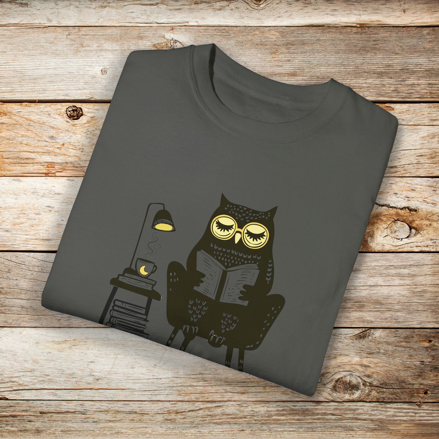 Medium gray Blue t-shirt featuring a line art drawing of an owl with large yellow eyes, sitting on a chair, reading a book. There is a stack of books beside her, and below, the text Night Owl is written in bold letters, adding a playful touch to the design.