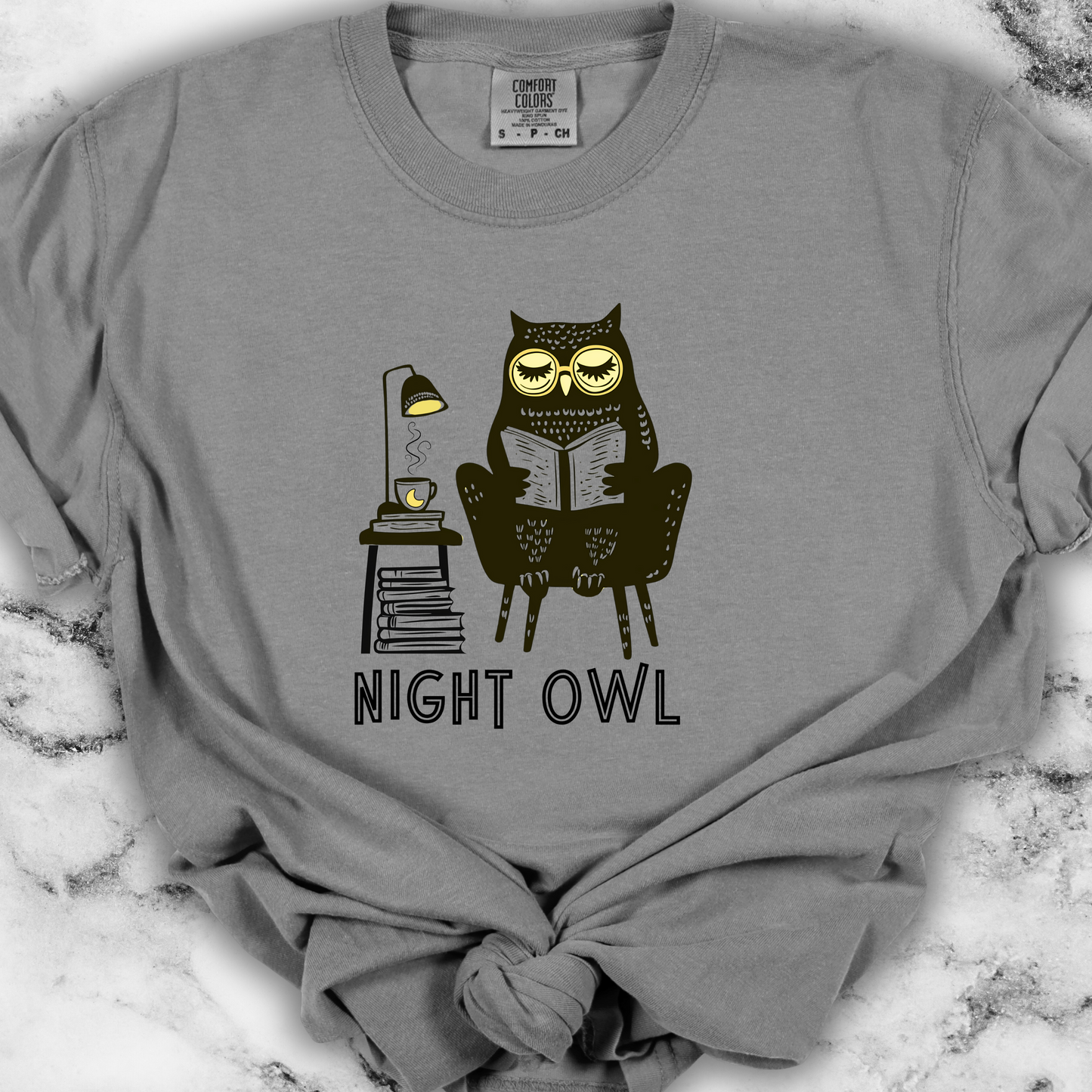 Light gray t-shirt featuring a line art drawing of an owl with large yellow eyes, sitting on a chair, reading a book. There is a stack of books beside her, and below, the text 'Night Owl' is written in bold letters, adding a playful touch to the design.