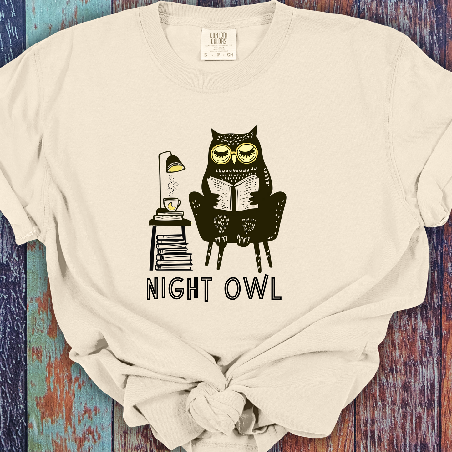 Ivory t-shirt featuring a line art drawing of an owl with large yellow eyes, sitting on a chair, reading a book. There is a stack of books beside her, and below, the text 'Night Owl' is written in bold letters, adding a playful touch to the design.