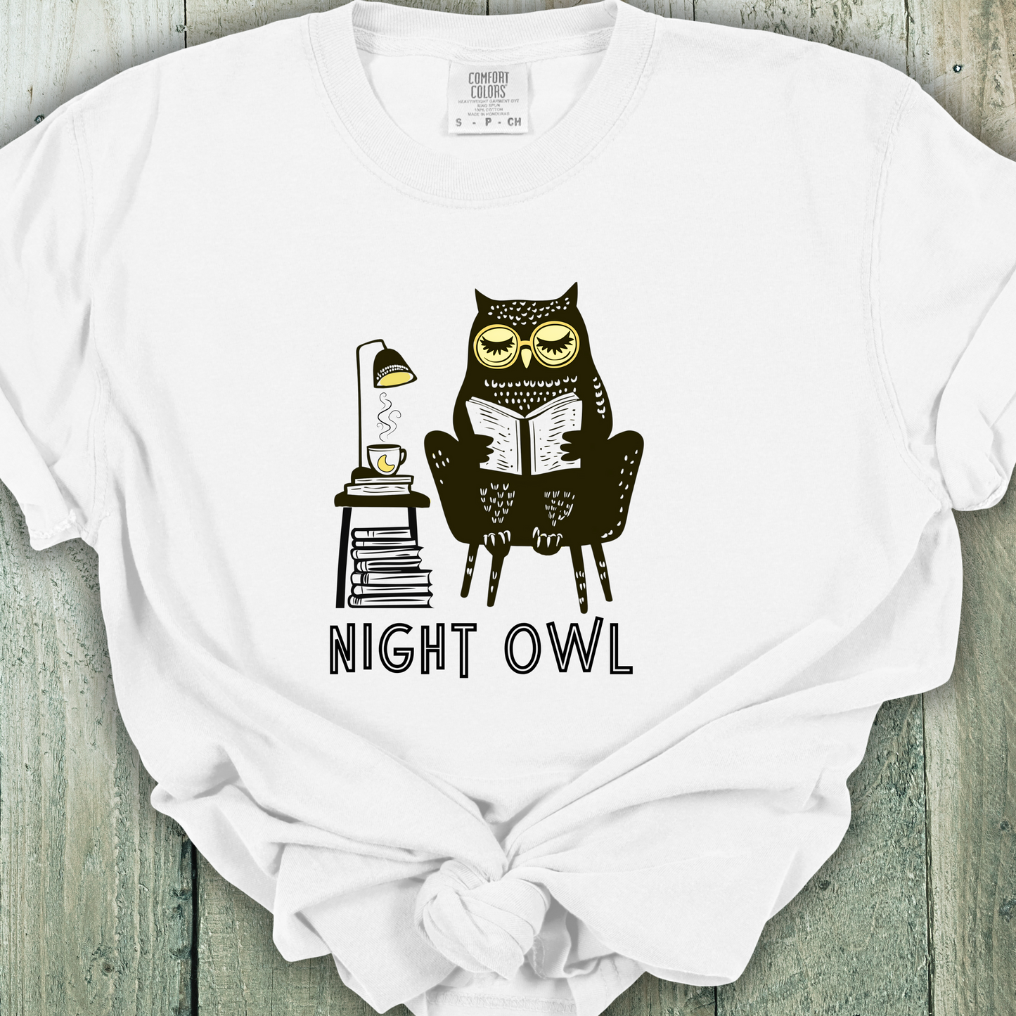 White t-shirt featuring a line art drawing of an owl with large yellow eyes, sitting on a chair, reading a book. There is a stack of books beside her, and below, the text 'Night Owl' is written in bold letters, adding a playful touch to the design.