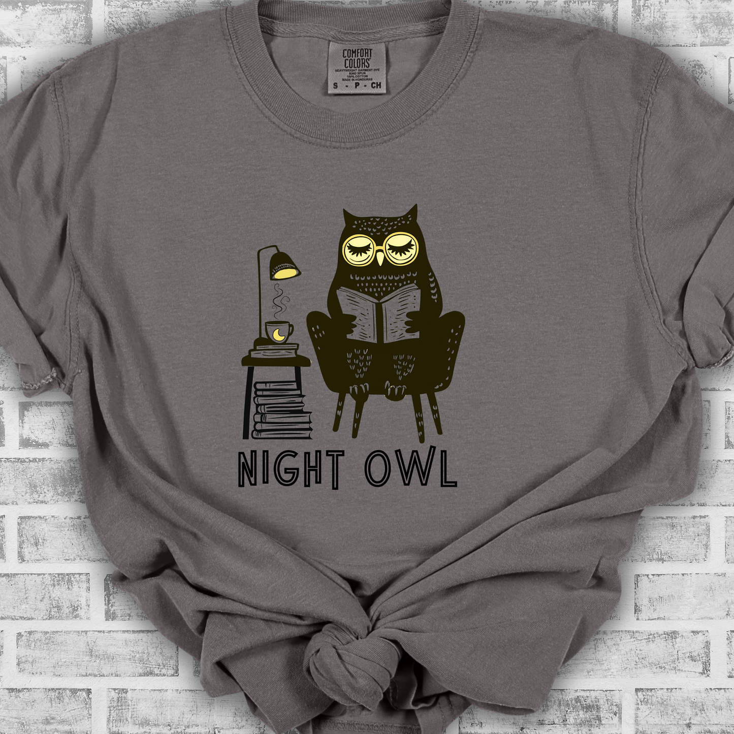 Medium gray t-shirt featuring a line art drawing of an owl with large yellow eyes, sitting on a chair, reading a book. There is a stack of books beside her, and below, the text 'Night Owl' is written in bold letters, adding a playful touch to the design.