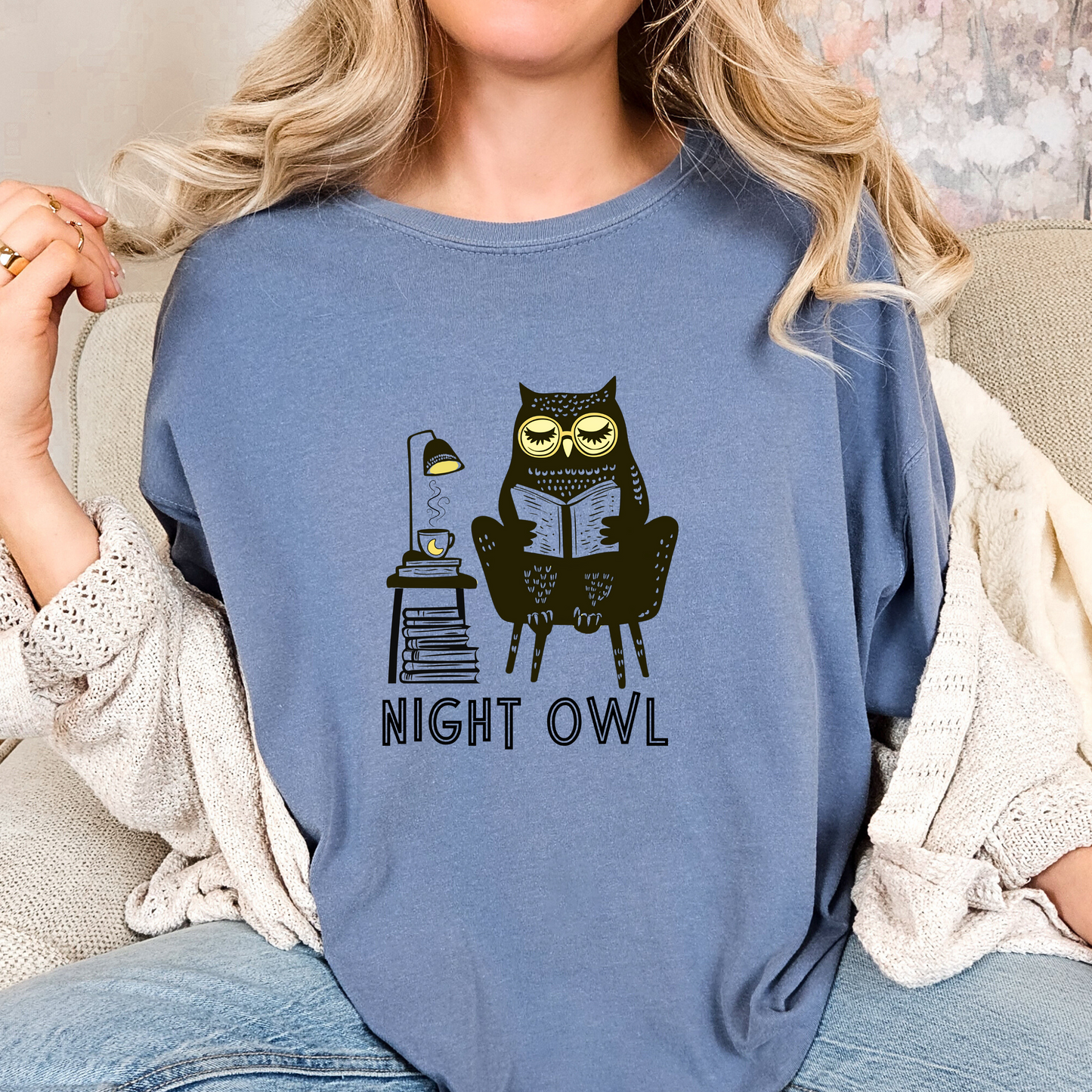 Blue t-shirt featuring a line art drawing of an owl with large yellow eyes, sitting on a chair, reading a book. There is a stack of books beside her, and below, the text 'Night Owl' is written in bold letters, adding a playful touch to the design.
