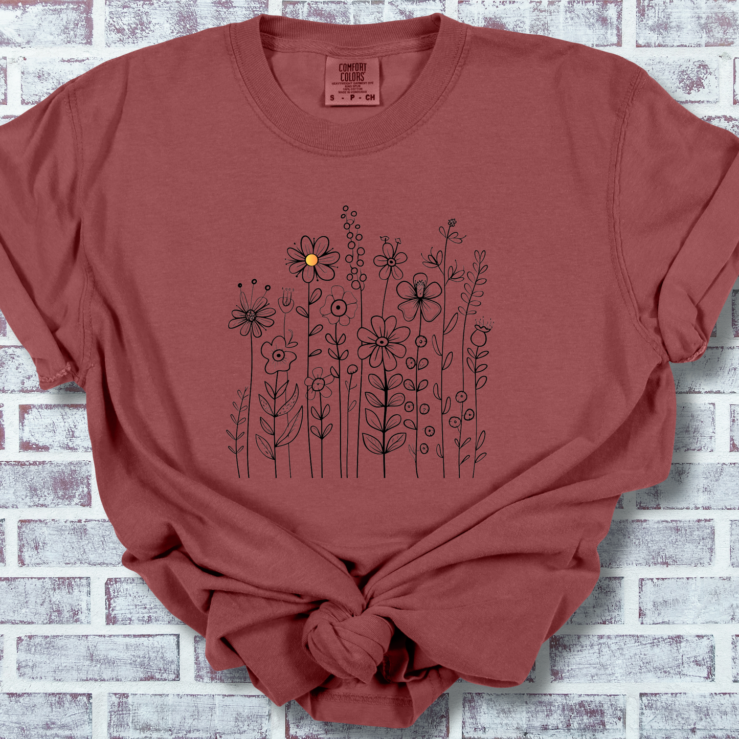 Cinnamon colored shirt with a minimalist black line art design featuring a variety of whimsical flowers and stems. One flower stands out with a small pop of yellow at its center, adding a subtle touch of color to the elegant floral arrangement.