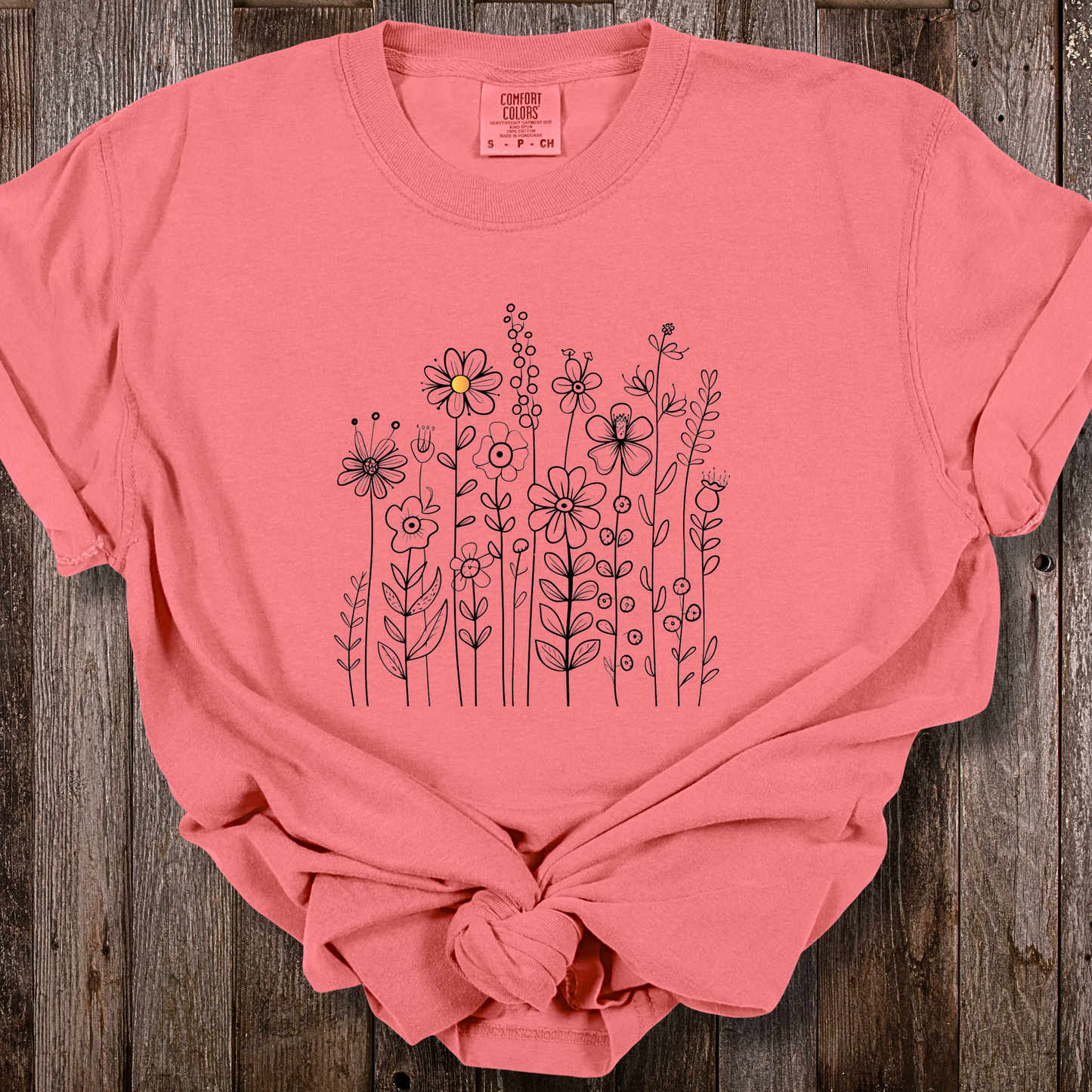 Watermelon pink shirt with a minimalist black line art design featuring a variety of whimsical flowers and stems. One flower stands out with a small pop of yellow at its center, adding a subtle touch of color to the elegant floral arrangement.