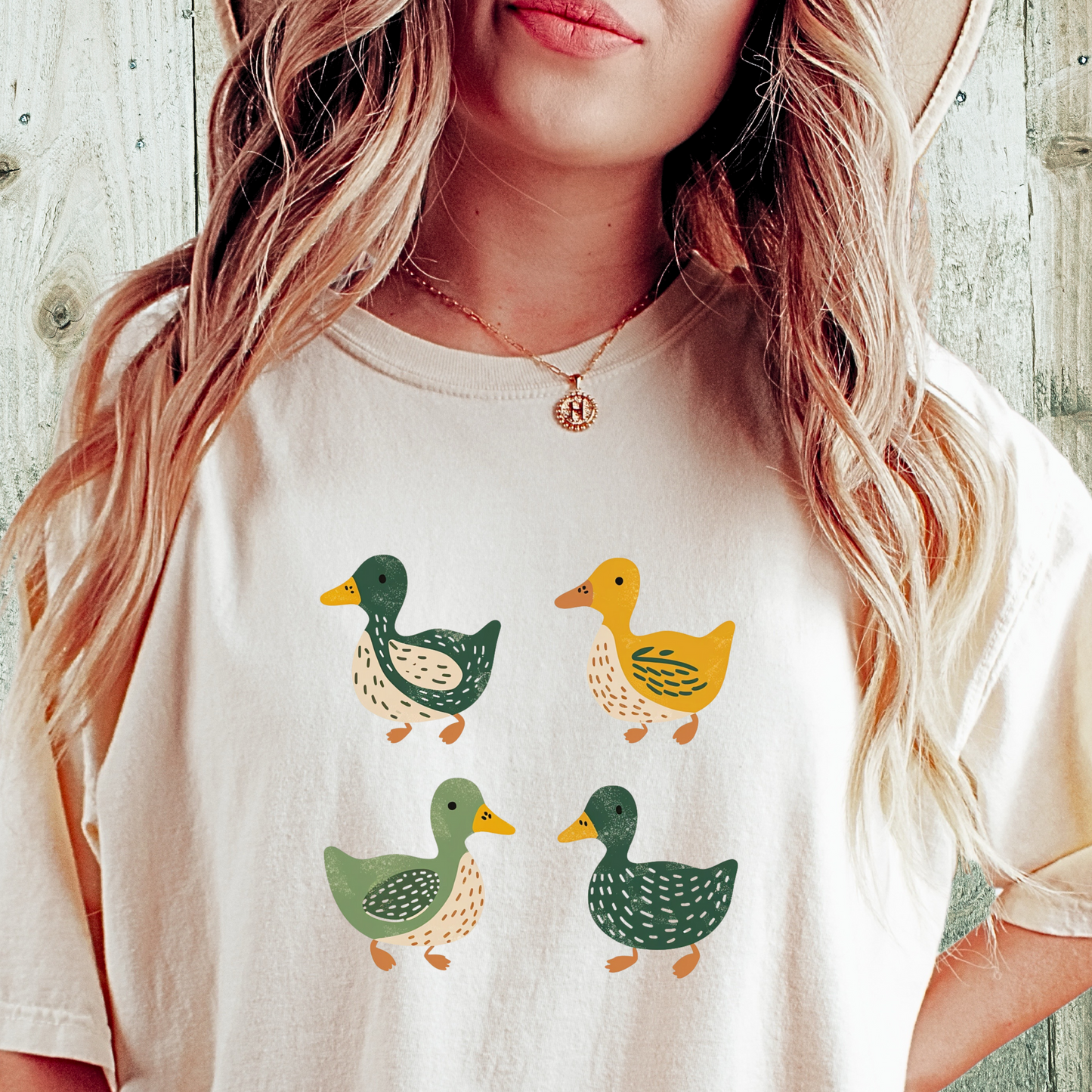Ivory t-shirt featuring a cute illustration of four cartoon ducks in earthy tones of green, yellow, and beige. Each duck has a unique color and pattern, arranged in a simple, playful style, adding a charming touch to the design.