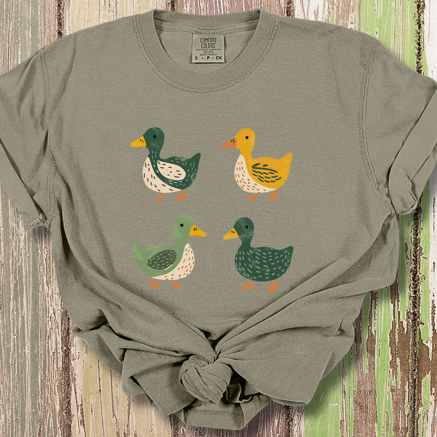 Khaki colored t-shirt featuring a cute illustration of four cartoon ducks in earthy tones of green, yellow, and beige. Each duck has a unique color and pattern, arranged in a simple, playful style, adding a charming touch to the design.