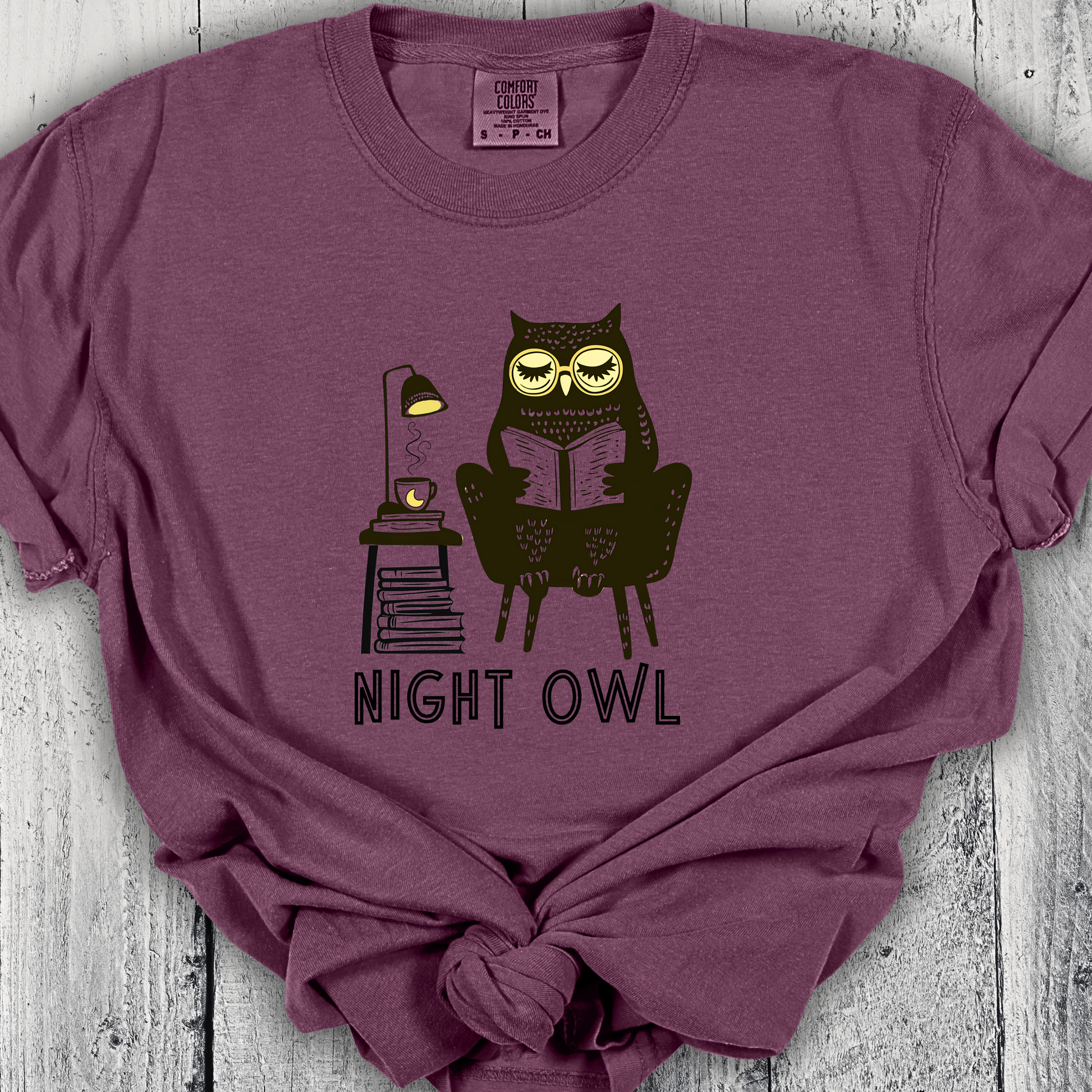 Purple t-shirt featuring a line art drawing of an owl with large yellow eyes, sitting on a chair, reading a book. There is a stack of books beside her, and below, the text &#39;Night Owl&#39; is written in bold letters, adding a playful touch to the design.