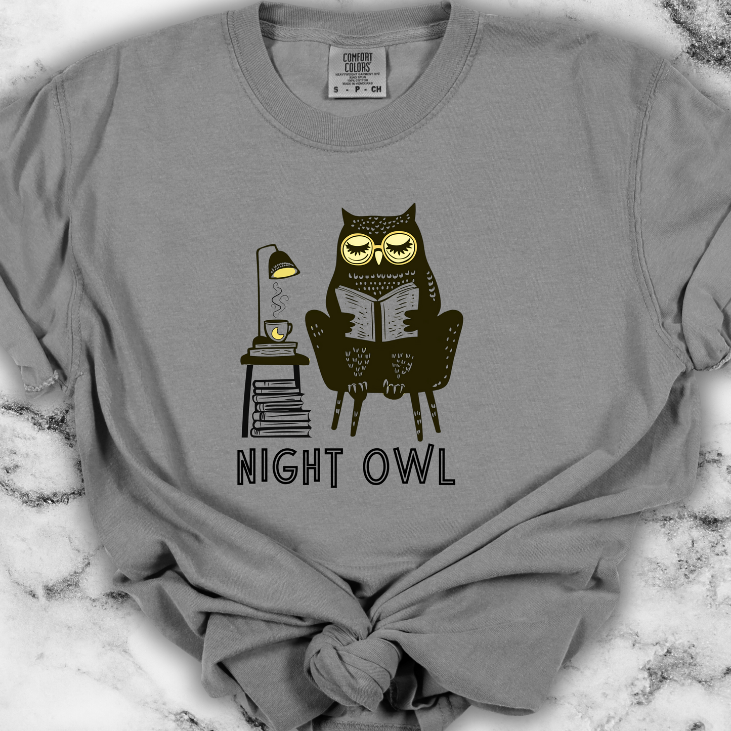 Light gray t-shirt featuring a line art drawing of an owl with large yellow eyes, sitting on a chair, reading a book. There is a stack of books beside her, and below, the text &#39;Night Owl&#39; is written in bold letters, adding a playful touch to the design.