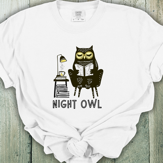 White  t-shirt featuring a line art drawing of an owl with large yellow eyes, sitting on a chair, reading a book. There is a stack of books beside her, and below, the text &#39;Night Owl&#39; is written in bold letters, adding a playful touch to the design.