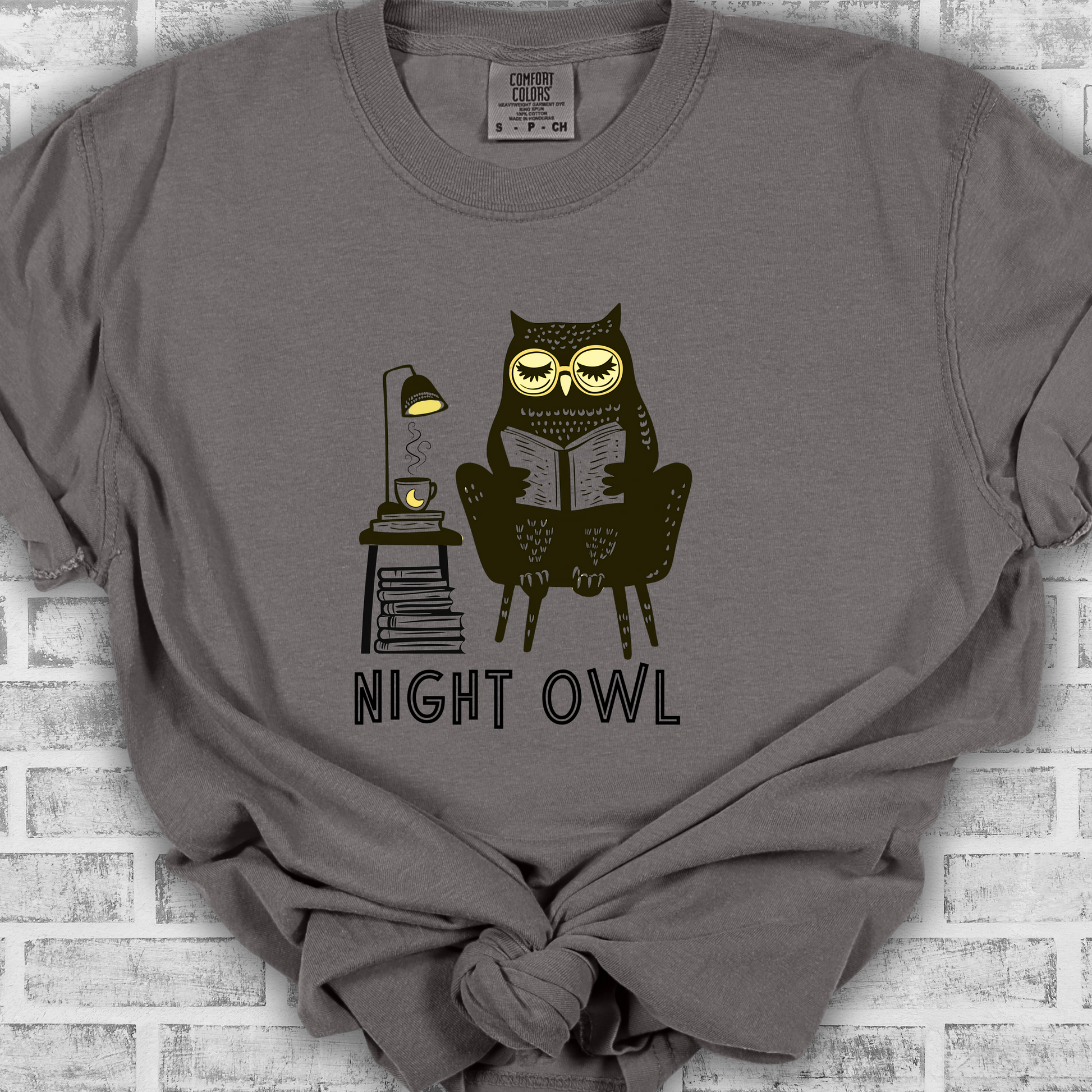 Medium gray t-shirt featuring a line art drawing of an owl with large yellow eyes, sitting on a chair, reading a book. There is a stack of books beside her, and below, the text &#39;Night Owl&#39; is written in bold letters, adding a playful touch to the design.