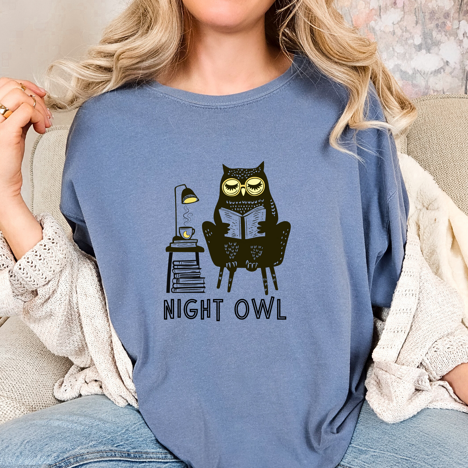 Blue t-shirt featuring a line art drawing of an owl with large yellow eyes, sitting on a chair, reading a book. There is a stack of books beside her, and below, the text &#39;Night Owl&#39; is written in bold letters, adding a playful touch to the design.