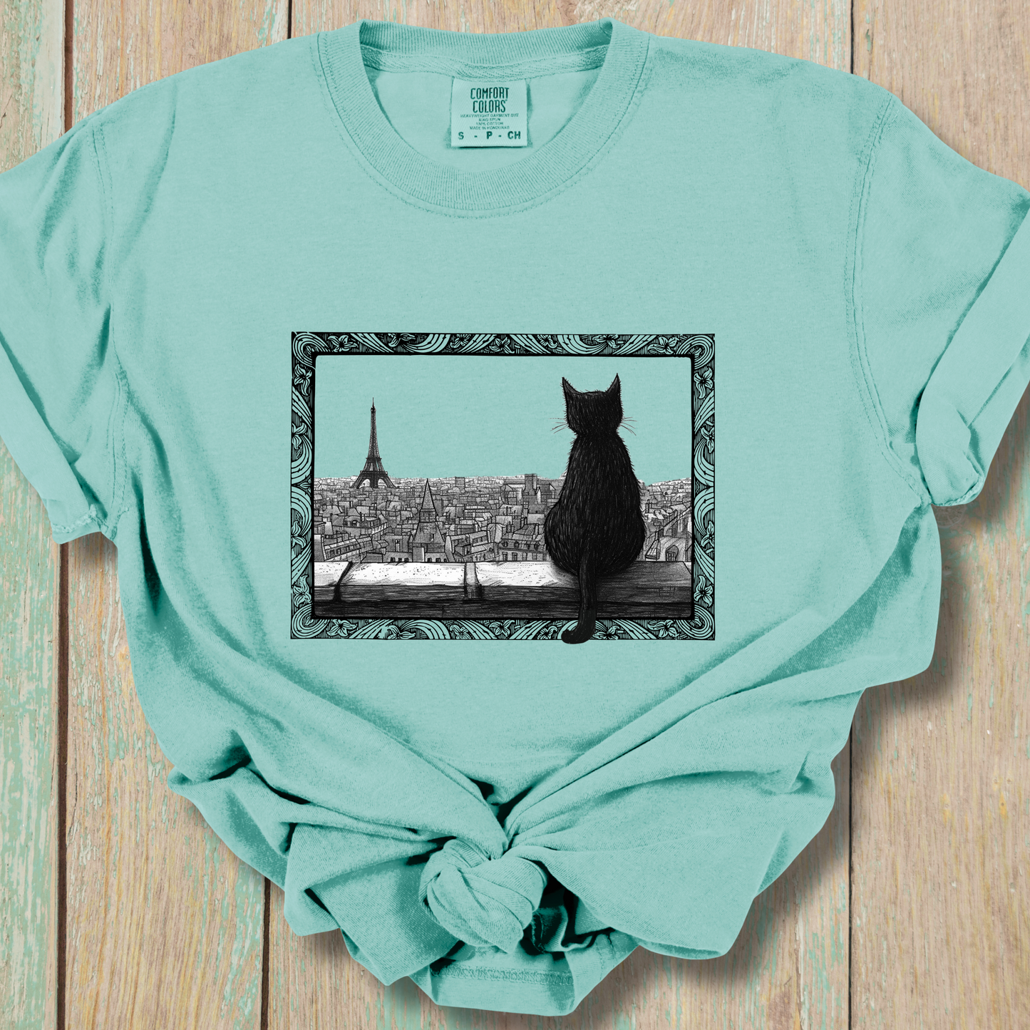 Light turquoise t-shirt featuring a black cat sitting on a windowsill, looking out over a cityscape with the Eiffel Tower in the background. The image is framed with intricate, decorative line patterns, adding an artistic and whimsical feel to the shirt.