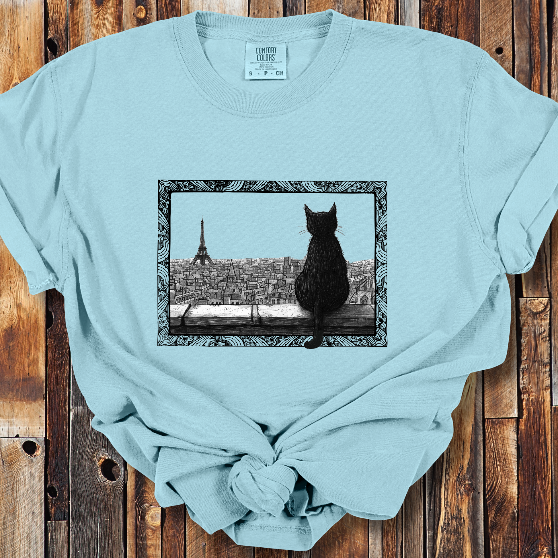 Chambray t-shirt featuring a black cat sitting on a windowsill, looking out over a cityscape with the Eiffel Tower in the background. The image is framed with intricate, decorative line patterns, adding an artistic and whimsical feel to the shirt.