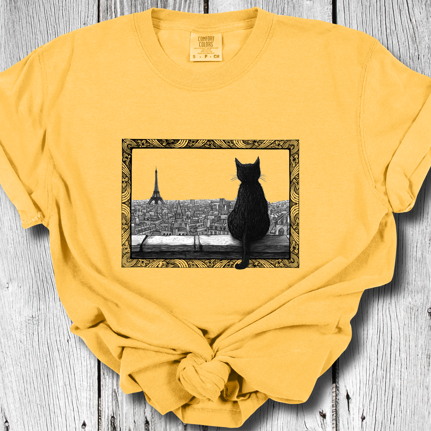 Citrus color t-shirt featuring a black cat sitting on a windowsill, looking out over a cityscape with the Eiffel Tower in the background. The image is framed with intricate, decorative line patterns, adding an artistic and whimsical feel.