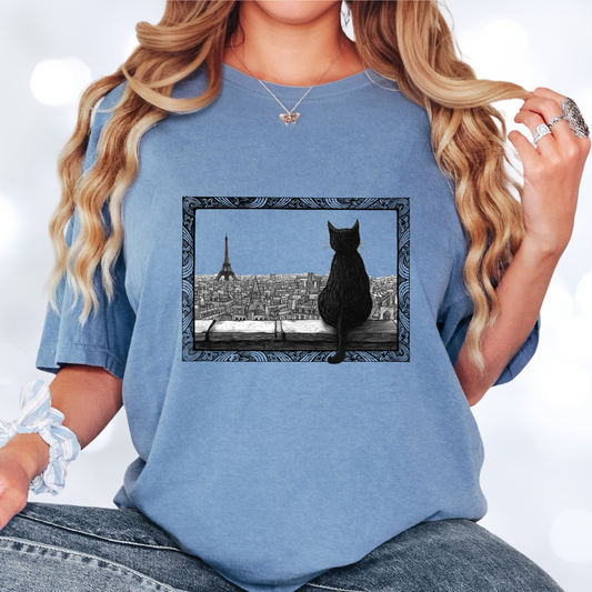 Blue jean colored t-shirt featuring a black cat sitting on a windowsill, looking out over a cityscape with the Eiffel Tower in the background. The image is framed with intricate, decorative line patterns, adding an artistic and whimsical feel.