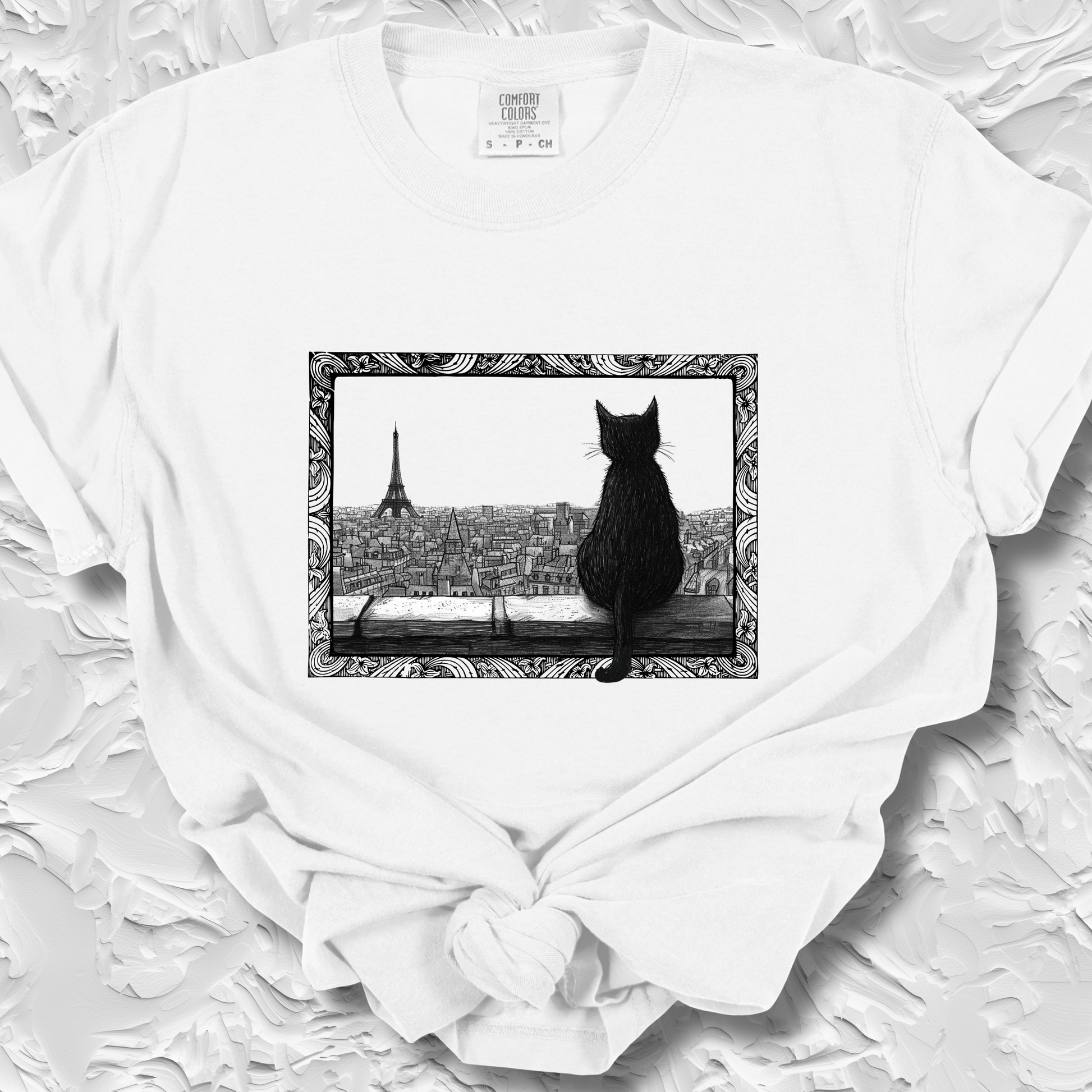 White t-shirt featuring a black cat sitting on a windowsill, looking out over a cityscape with the Eiffel Tower in the background. The image is framed with intricate, decorative line patterns, adding an artistic and whimsical feel.