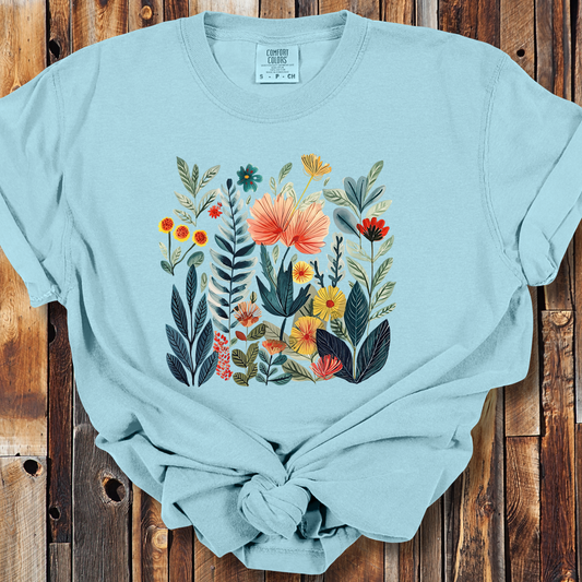 Light blue t-shirt featuring a vibrant, detailed illustration of various flowers and foliage in a cottage garden style. The design includes large blooms in shades of coral, yellow, and red, surrounded by lush green leaves and smaller flowers, creating a colorful and lively floral arrangement.