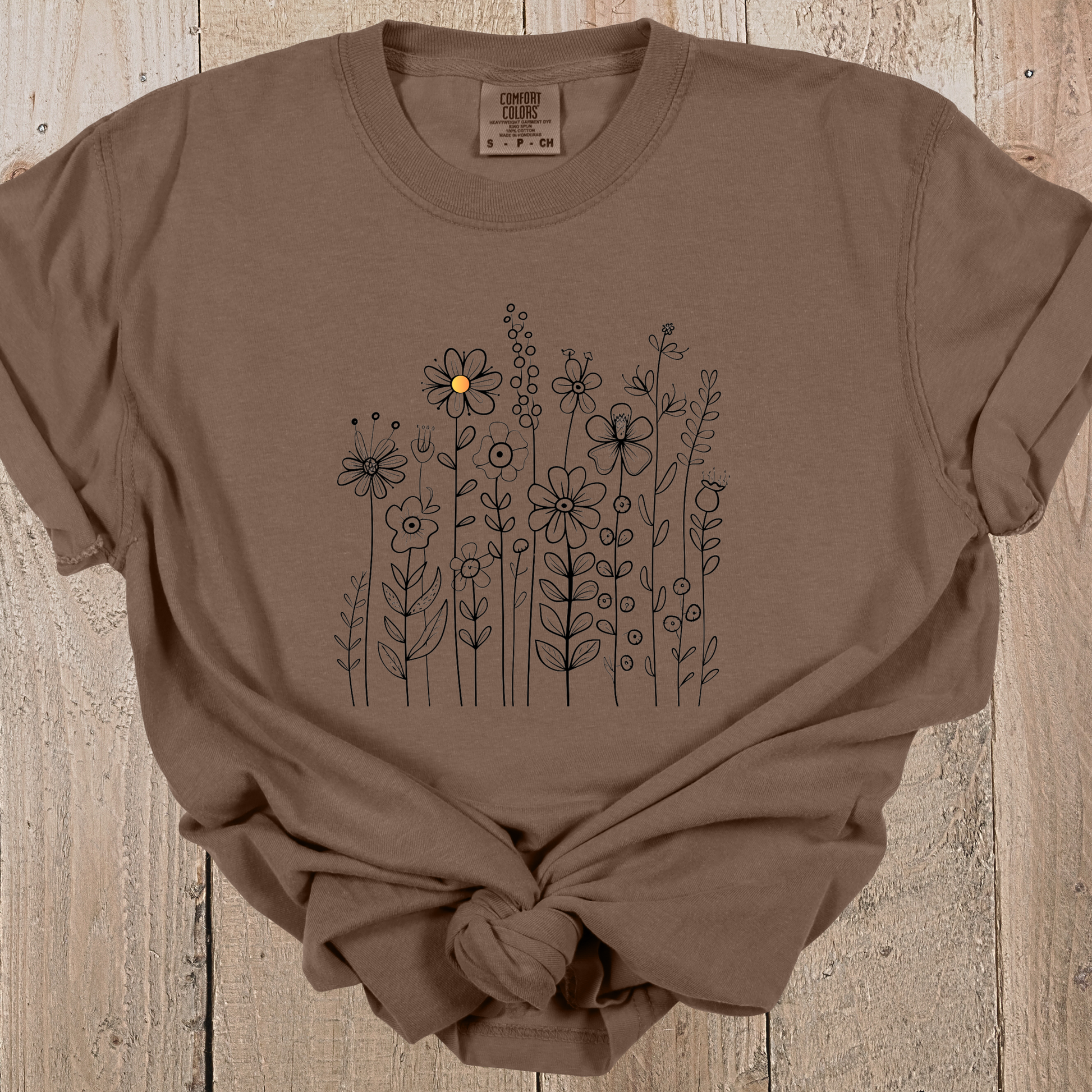 Espresso colored t-shirt with a minimalist black line art design featuring a variety of whimsical flowers and stems. One flower stands out with a small pop of yellow at its center, adding a subtle touch of color to the elegant floral arrangement.